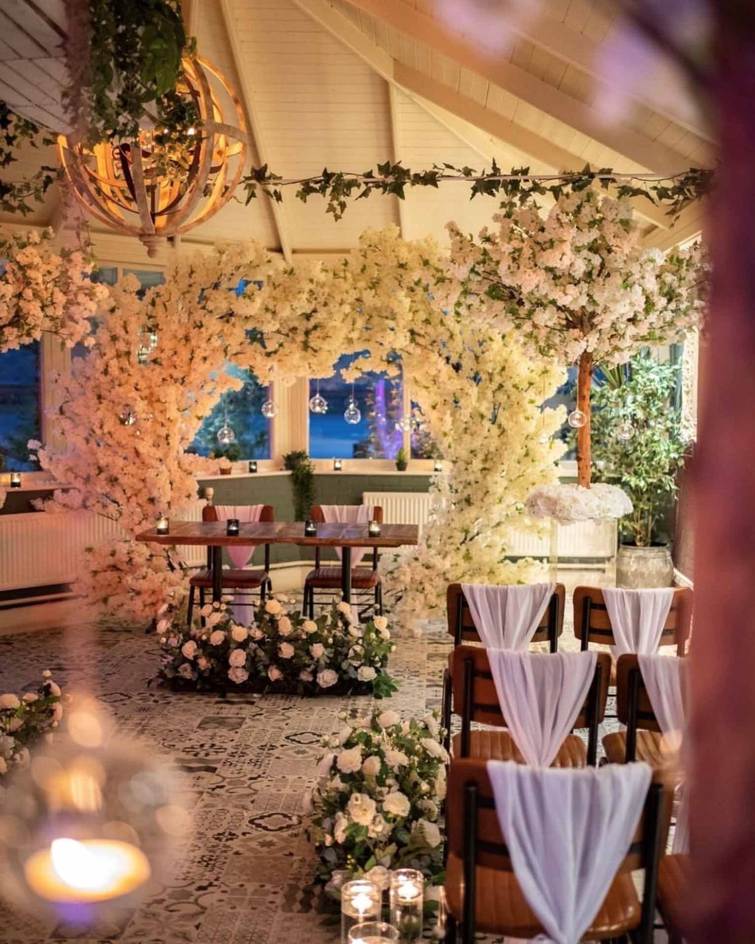 A wedding ceremony set beneath an arch of white blossom, with candles and draped chairs